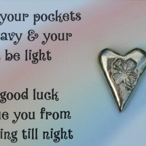 May include: A silver heart charm with a four-leaf clover design. The charm is on a card with the text "May your pockets be heavy & your heart be light. May good luck persue you from morning till night."