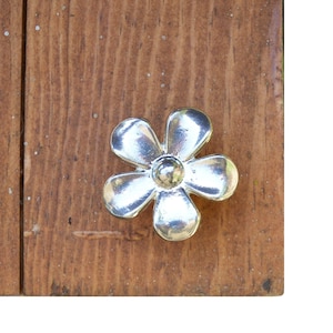 May include: A silver flower-shaped cabinet knob with five petals. The knob is mounted on a wooden surface.