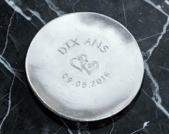 Op de afbeelding: Een ronde, zilverkleurige metalen schaal met een glad, reflecterend oppervlak. In het midden is de tekst DIX ANS, twee in elkaar grijpende hartvormen en de numerieke datum 09.05.2016 gestempeld in een eenvoudig, schreefloos lettertype.