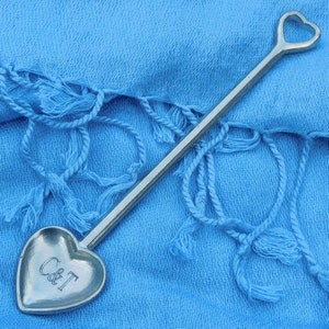 Könnte beinhalten: Ein silberner herzförmiger Löffel mit einem Griff und einer herzförmigen Schale. Die Schale ist mit den Buchstaben "C & T" graviert. Der Löffel liegt auf einem hellblauen Stoff mit Fransen.