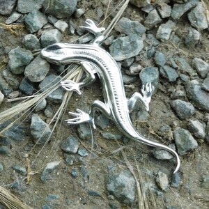Puede incluir: Una figura de lagarto plateado con escamas detalladas y extremidades articuladas. El lagarto est&aacute; sobre un lecho de peque&ntilde;as rocas grises y tierra, con algunos mechones de hierba seca. La figura tiene una superficie brillante y reflectante.