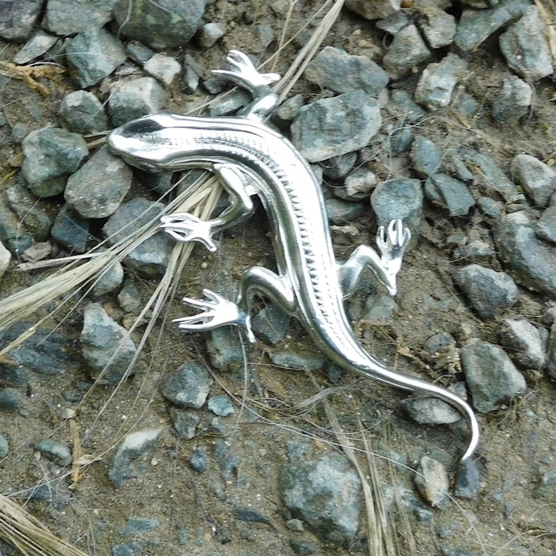 Puede incluir: Una figura de lagarto plateado con escamas detalladas y extremidades articuladas. El lagarto est&aacute; sobre un lecho de peque&ntilde;as rocas grises y tierra, con algunos mechones de hierba seca. La figura tiene una superficie brillante y reflectante.