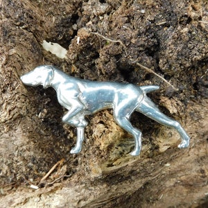 Könnte beinhalten: Eine silberfarbene Metallfigur eines Hundes in Laufpose. Der Hund ist ein Pointer, mit einem schlanken, detaillierten Design. Die Figur befindet sich auf einer strukturierten, braunen Holzoberfläche.