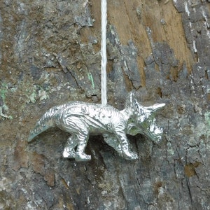 May include: A silver-coloured, detailed, toy triceratops dinosaur hanging from a white string. The dinosaur is facing the right side of the image.