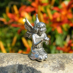 Peut inclure: Une figurine en argent d'une fée aux ailes déployées, agenouillée, les mains jointes en prière. La fée porte une robe et a un visage délicat.