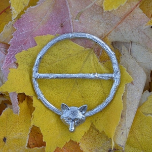 May include: A silver ring featuring a fox head design, set against a background of yellow and pink autumn leaves.