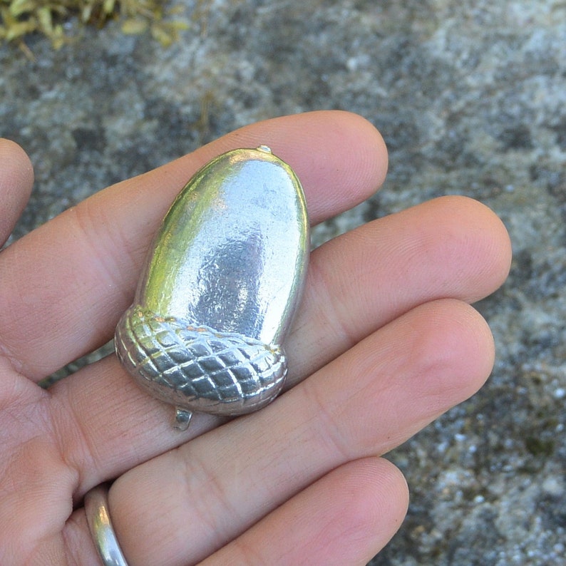 May include: A silver acorn charm with a detailed, textured cap. The charm is smooth and shiny, with a slightly rounded shape.