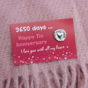May include: A red card with the text "3650 days... Happy Tin Anniversary I love you with all my heart x" and a silver heart-shaped pin. The card is decorated with small heart illustrations and rests on a pink, textured surface.