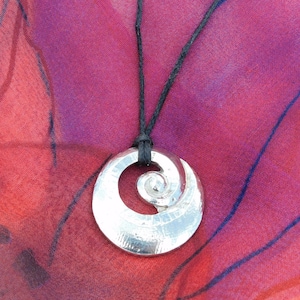 Pode incluir: Um colar com um pingente de prata com um design em espiral pendurado em um cordão preto sobre um fundo vermelho e roxo.