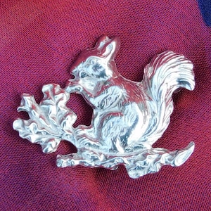 May include: A silver squirrel brooch with a detailed design, sitting on a branch with leaves. The squirrel is holding a nut in its paws.