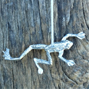 以下が含まれることがあります： 白い紐に吊るされた銀色のカエルの置物。カエルは足を伸ばした跳躍のポーズをとっている。置物は、質感のある鱗で細部まで作られており、背面には吊り下げ用の穴が開いている。