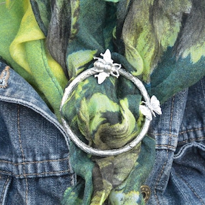 Pode incluir: Um anel de prata com um design de abelha e borboleta em um lenço estampado em verde e amarelo. O lenço está drapeado sobre uma jaqueta de ganga.