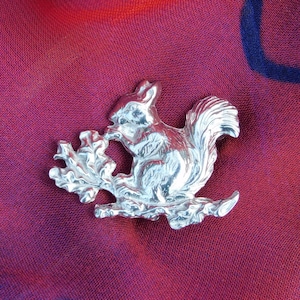 May include: A silver squirrel brooch with a detailed design of the squirrel perched on a branch with leaves. The brooch is a delicate and intricate piece of jewelry.