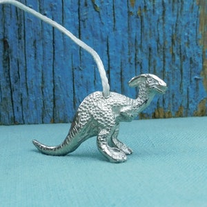 Pode incluir: Um amuleto de dinossauro prateado com uma superfície texturizada. O amuleto está pendurado em um cordão branco.