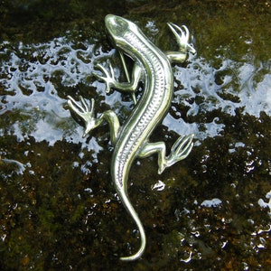 Puede incluir: Broche plateado con forma de lagarto, con escamas detalladas y extremidades extendidas. El alfiler con forma de reptil se encuentra sobre un fondo oscuro y h&uacute;medo, creando un contraste llamativo. El broche tiene un cierre de alfiler.
