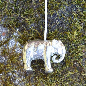 Peut inclure: Un pendentif éléphant argenté avec une trompe recourbée et des détails fins est suspendu à une ficelle blanche. Le pendentif est posé sur un fond de mousse verte et brune, créant un contraste naturel.