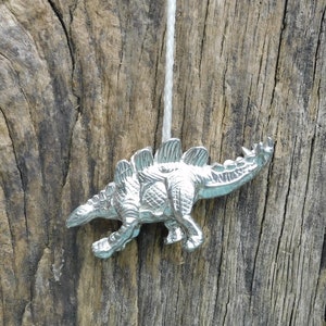 Può includere: Un ciondolo a forma di dinosauro color argento appeso a un cordoncino bianco. Il dinosauro è rivolto a destra e ha una schiena spinosa.