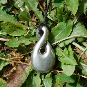 Può includere: Un ciondolo d'argento a forma di simbolo di infinito stilizzato, appeso a un cordoncino nero. Il ciondolo è appoggiato su un letto di foglie verdi.