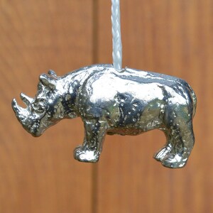 May include: A silver-coloured rhinoceros figurine hanging from a white cord. The figurine is detailed with textured skin and a curled horn.