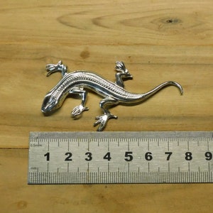 Puede incluir: Una detallada figura de lagarto plateado, de aproximadamente 10 cm de largo, con intrincados detalles en su cuerpo y patas. El lagarto est&aacute; posicionado sobre una superficie de madera, mostrando su dise&ntilde;o realista. La cola de la figura se curva con gracia.