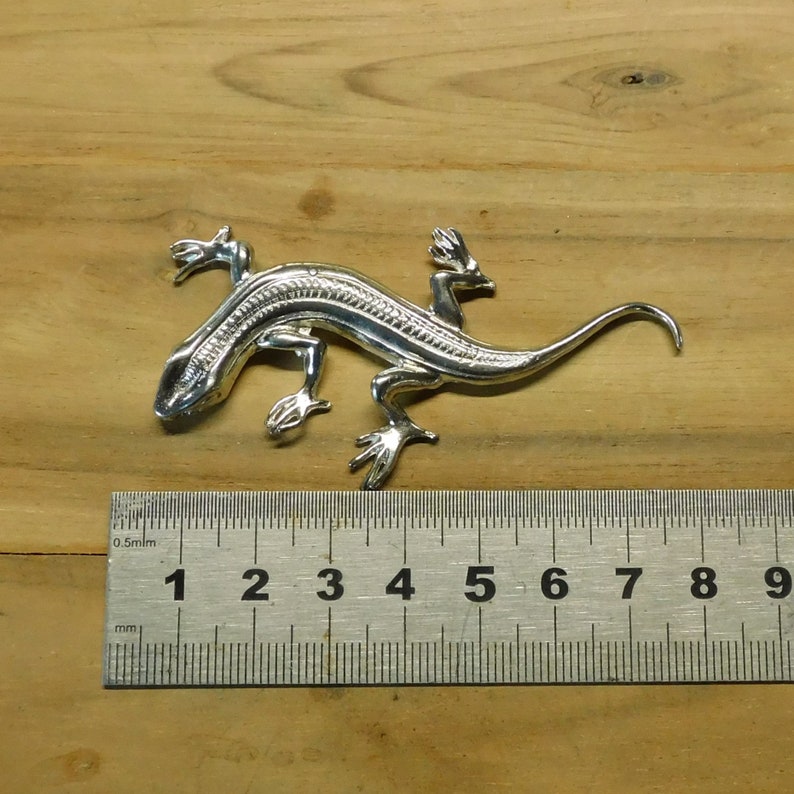 Puede incluir: Una detallada figura de lagarto plateado, de aproximadamente 10 cm de largo, con intrincados detalles en su cuerpo y patas. El lagarto est&aacute; posicionado sobre una superficie de madera, mostrando su dise&ntilde;o realista. La cola de la figura se curva con gracia.
