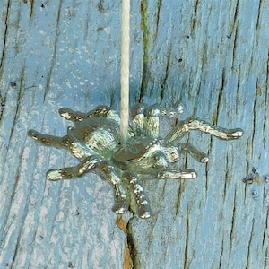 Può includere: Una statuetta di ragno color argento appesa a uno spago bianco. Il ragno ha otto zampe e un corpo dettagliato.