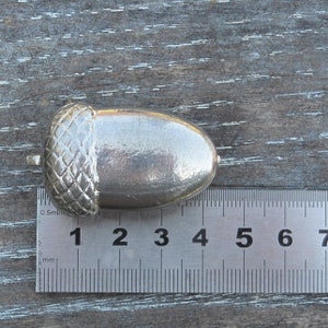 May include: A silver acorn charm with a textured cap, measuring approximately 3 cm in length. The charm is shown next to a ruler.