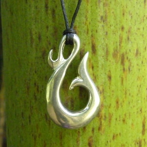 May include: A silver pendant in the shape of a fish hook hangs from a black cord. The pendant is shiny and has a detailed design. The pendant is hanging against a green background.