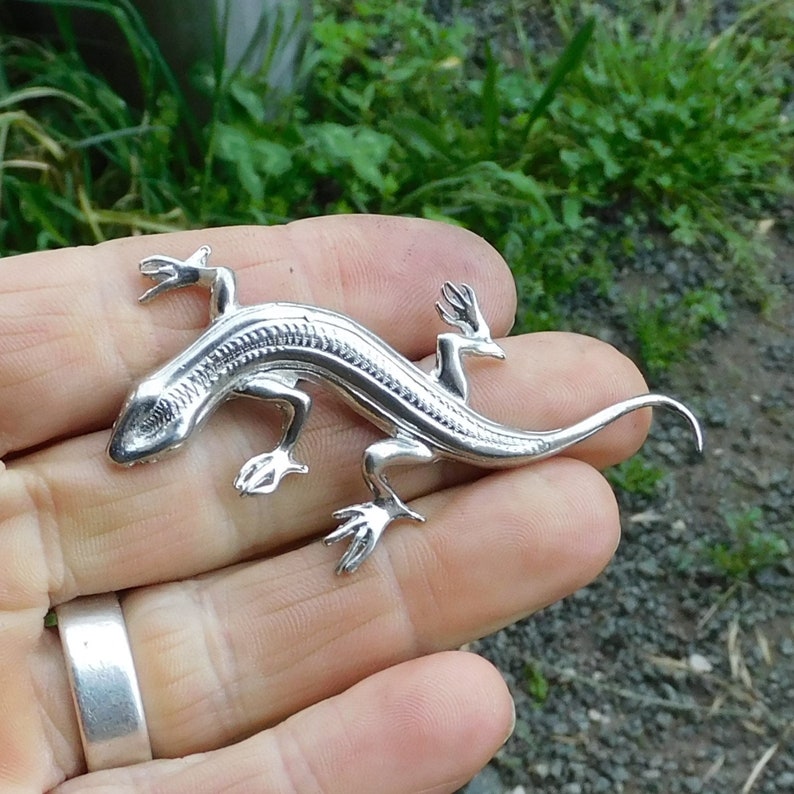 Puede incluir: Una detallada figura de lagarto plateado con cuatro patas, una larga cola y un cuerpo texturizado. El lagarto se sostiene en una mano sobre un fondo de follaje verde.