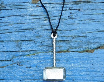 Op de afbeelding: Een zilverkleurige hamerhanger aan een zwarte koordsnoer. De hanger is een replica van Thors hamer, Mjolnir, uit de Noorse mythologie.