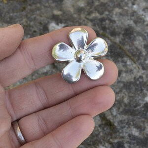 May include: A silver flower charm with five petals and a round centre. The charm is made of metal and has a shiny finish.