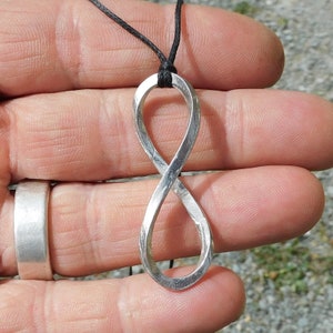 Peut inclure: Un collier avec un pendentif en forme d'infini en argent sur un cordon noir. Le pendentif est en métal et a une finition lisse et polie.