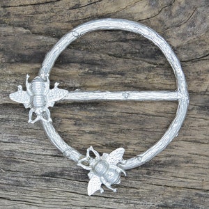 Pode incluir: Fecho de botão de metal prateado em forma de abelha com um design de galho. O fecho tem a forma de um círculo com uma barra no centro. As abelhas são detalhadas com asas e pernas.