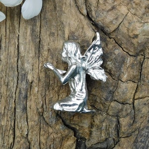 Peut inclure: Une figurine en argent d'une fée aux ailes déployées, agenouillée, les mains jointes en prière. La fée est sur un fond de bois rustique.