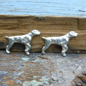 May include: Two silver-coloured metal figurines of German Shorthaired Pointers, standing on a wooden surface.