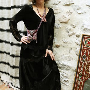 May include: A black long-sleeved dress featuring a V-neckline, lace trim on the sleeves and hem, and a layered skirt. A small, patterned crossbody bag with a beaded strap and a matching beaded necklace are worn over the dress.