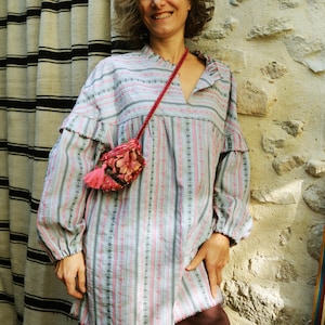 May include: A woman wearing a light blue and pink striped blouse with a pink and floral embroidered pouch hanging from a strap around her neck. The blouse has a V-neck and long sleeves.