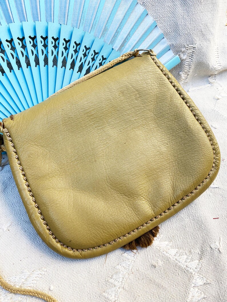 May include: A small, olive green leather purse with a fringe tassel. The purse has a simple design and a braided strap.