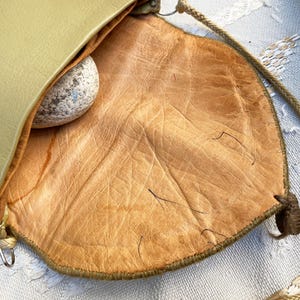 May include: A green leather bag with a brown leather lining. The bag has a long strap and a small pocket on the front.