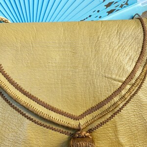 May include: A gold leather purse with a brown braided trim and a tassel. The purse has a flap closure and a top handle.