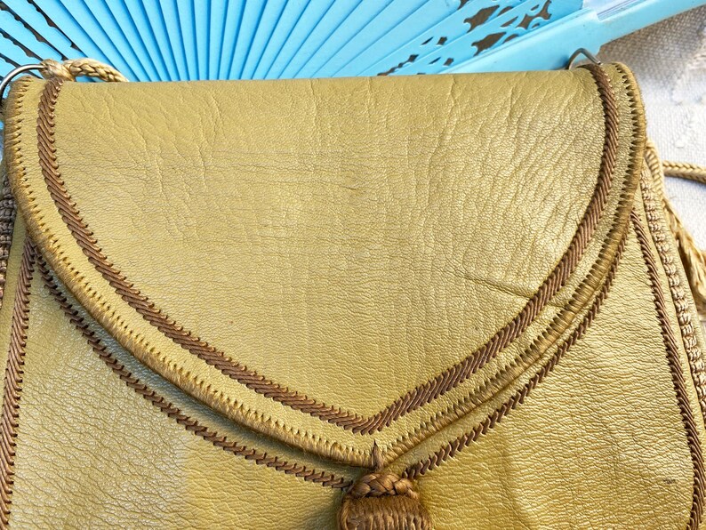 May include: A gold leather purse with a brown braided trim and a tassel. The purse has a flap closure and a top handle.