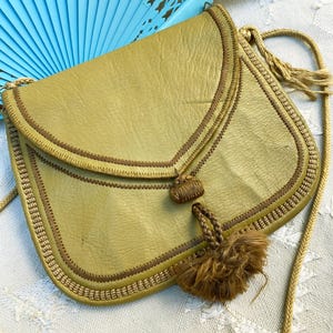 May include: A small, olive green leather purse with a brown tassel and a braided strap. The purse has a flap closure and is decorated with brown stitching.