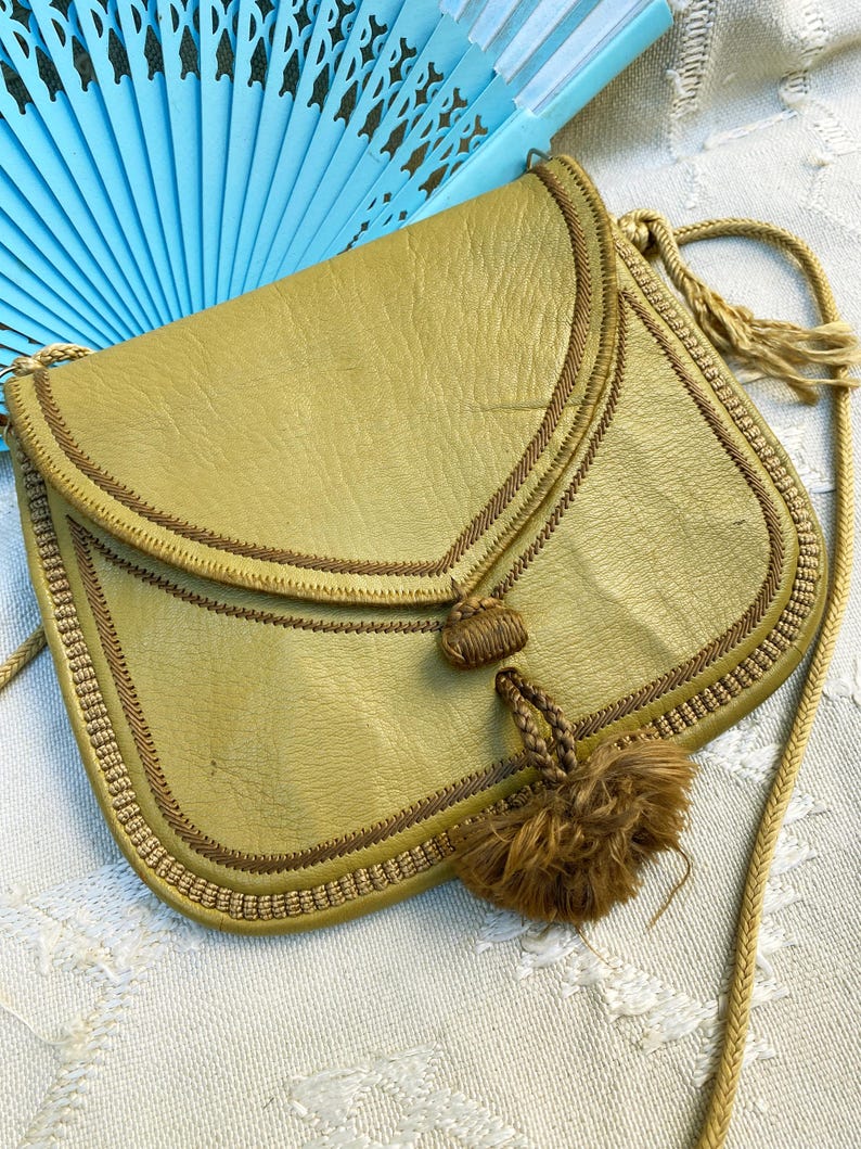 May include: A small, olive green leather purse with a brown tassel and a braided strap. The purse has a flap closure and is decorated with brown stitching.