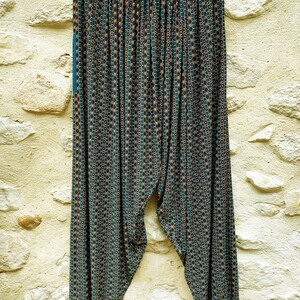 May include: A pair of green, black, and orange patterned pants with a wide waistband. The pants are hanging on a hanger against a stone wall.