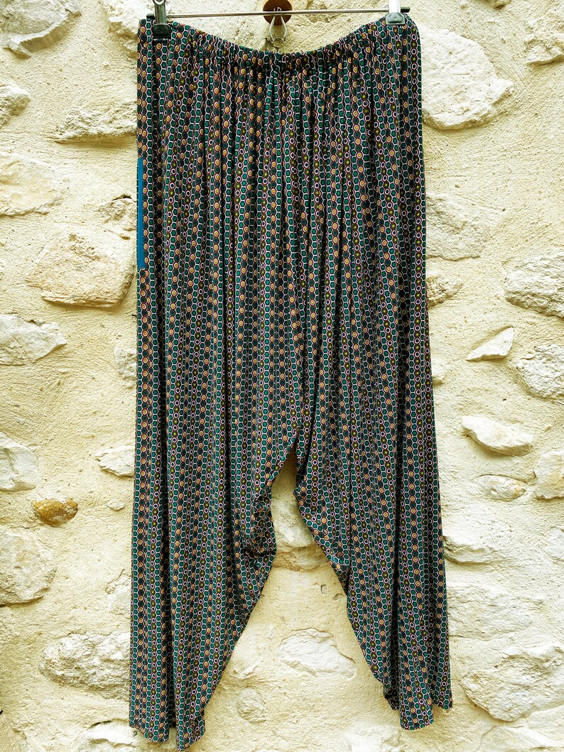 May include: A pair of green, black, and orange patterned pants with a wide waistband. The pants are hanging on a hanger against a stone wall.