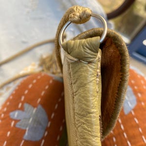 May include: A close-up of a light brown leather purse with a silver ring on the strap. The purse is sitting on a patterned fabric.