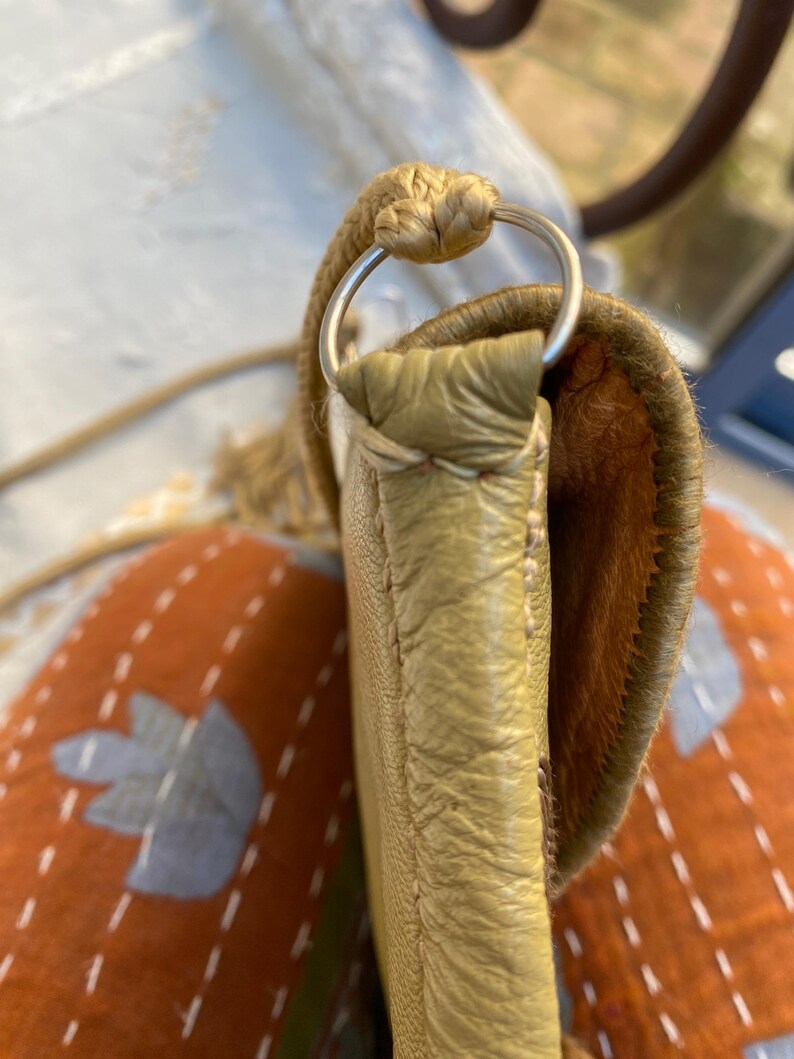 May include: A close-up of a light brown leather purse with a silver ring on the strap. The purse is sitting on a patterned fabric.
