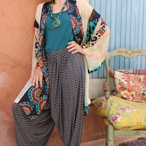 May include: A woman wearing a colorful patterned kimono with a teal tank top and patterned pants. She is standing in front of a blue wall and a chair with a floral patterned cushion.