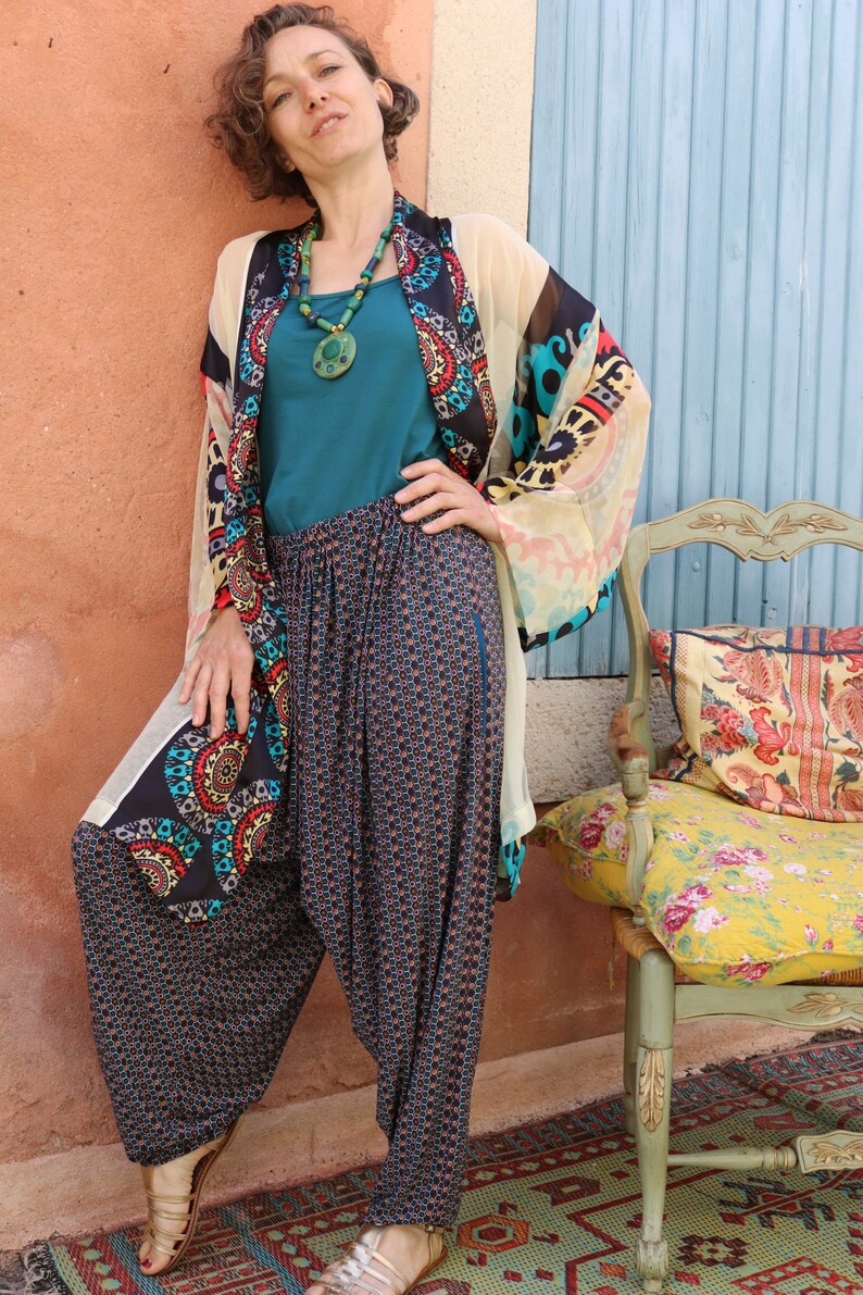 May include: A woman wearing a colorful patterned kimono with a teal tank top and patterned pants. She is standing in front of a blue wall and a chair with a floral patterned cushion.