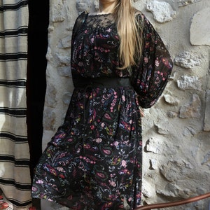 Puede incluir: Una mujer con un vestido negro floral con cintur&oacute;n negro y mangas largas. Est&aacute; de pie frente a una pared de piedra y tiene el pelo largo y rubio. Lleva gafas y una diadema.